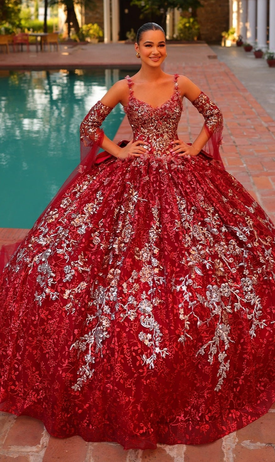 A young woman stands by a pool wearing the Quinceanera Dress 54322 By Amarra, a stunning floral embroidered ball gown in rich red with silver details and matching detached sleeves.